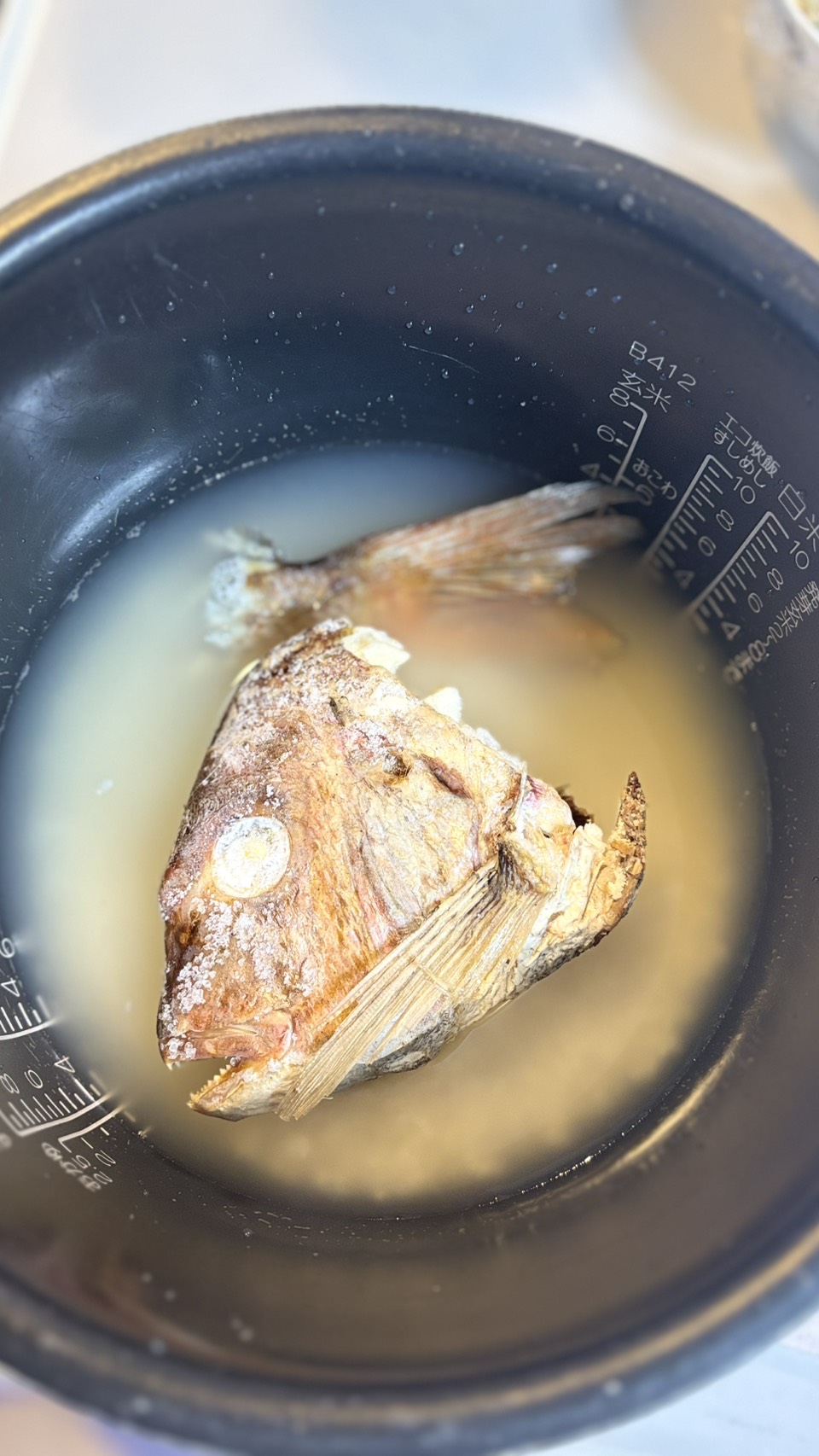 炊飯器にお米と黄金色のお出汁を入れ、冷凍した鯛のかぶと(頭)をのせたところ